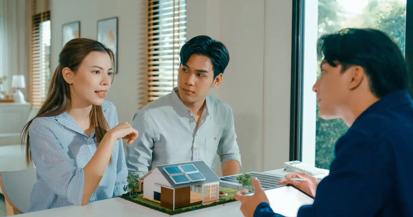 Três pessoas discutem planejamento de construção sustentável em uma reunião, com maquete de uma casa ecológica e painel solar, em ambiente bem iluminado.