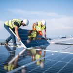 Dois técnicos instalando painéis solares em um telhado sob o céu claro, ilustrando as etapas práticas do processo de instalação de energia solar. Imagem destacando a importância de profissionais qualificados e equipamentos adequados para garantir eficiência e segurança no sistema fotovoltaico.