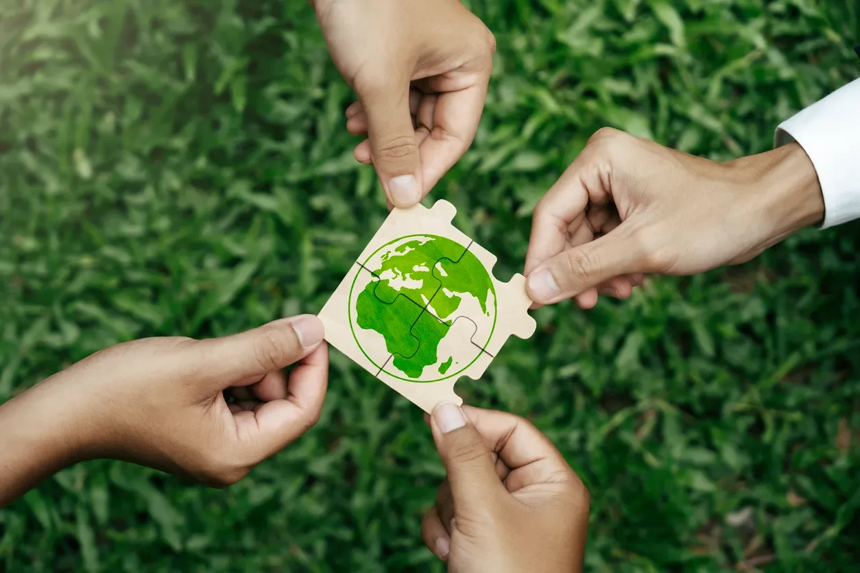 Diversas mãos unindo peças de um quebra-cabeça com um desenho de um planeta Terra, simbolizando sustentabilidade e preservação ambiental.