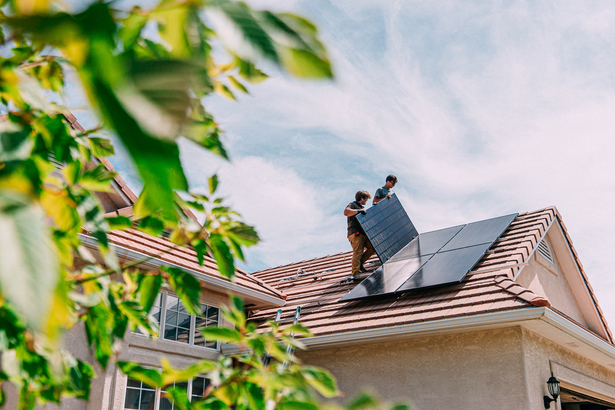 Dois profissionais instalando painéis solares no telhado, demonstrando práticas de sustentabilidade e eficiência energética em uma casa eco-friendly. A cena enfatiza a importância de energia limpa e inspirações para residências sustentáveis.