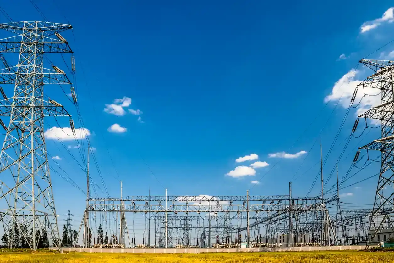 Imagem de uma subestação de energia com torres de transmissão de alta tensão sob céu azul com algumas nuvens, destacando a infraestrutura de transmissão de energia elétrica.