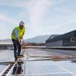 Técnico de limpeza em painel solar com equipamento de segurança, limpando as placas fotovoltaicas sob um céu azul.