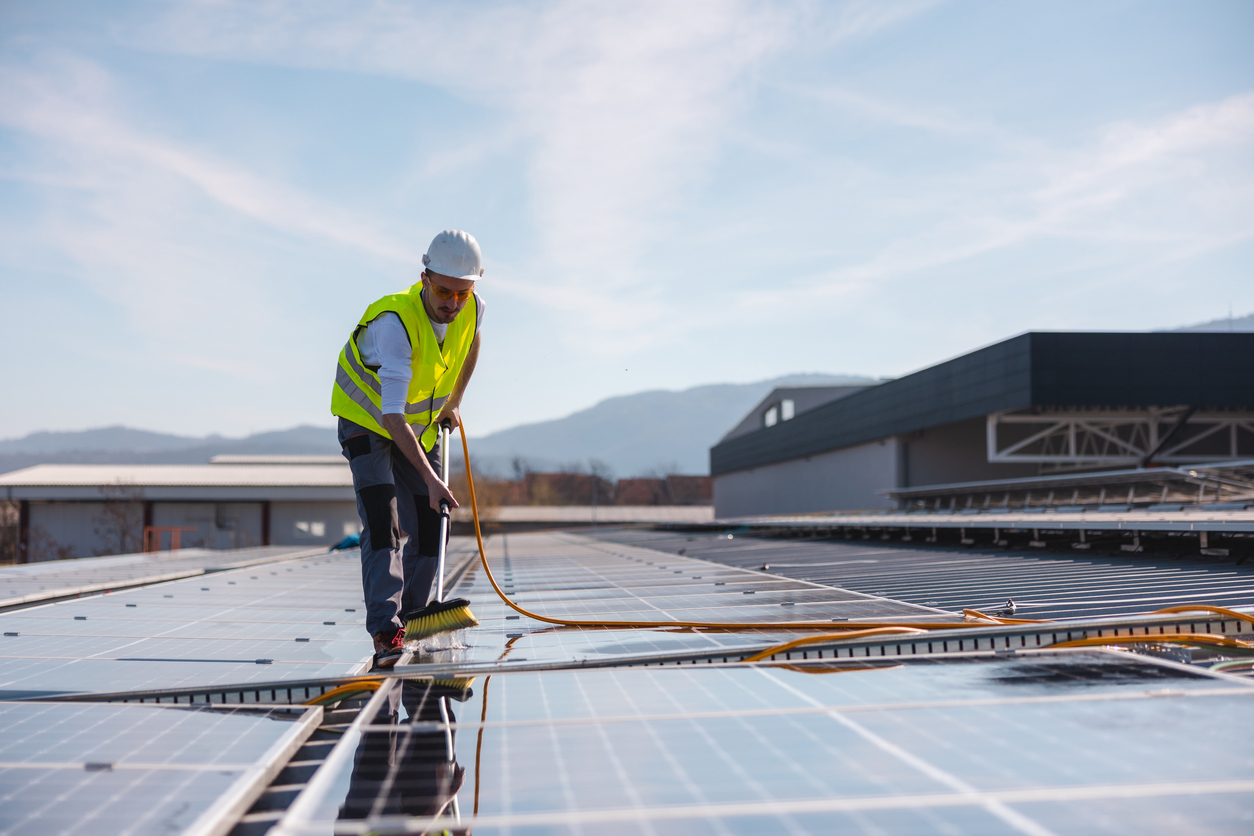 Técnico de limpeza em painel solar com equipamento de segurança, limpando as placas fotovoltaicas sob um céu azul.