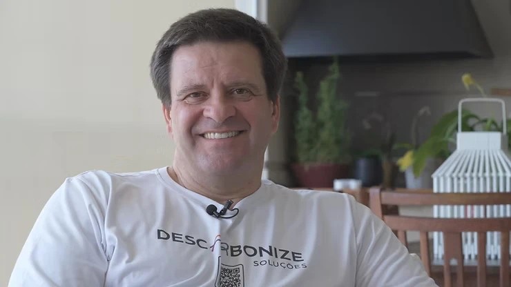 Homem sorridente usando camiseta da iniciativa Descarbonize Soluções. Ele aparece em um ambiente iluminado e tranquilo, cercado por plantas. A imagem transmite uma mensagem positiva sobre sustentabilidade e ações climáticas.