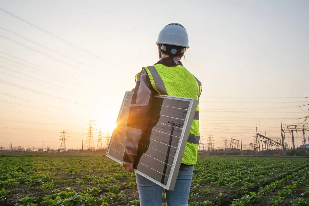Engenheira ou técnica de energia solar segurando um painel solar portátil em um campo agrícola durante o pôr do sol, com torres de energia e linhas de transmissão ao fundo, promovendo energia renovável e sustentável.