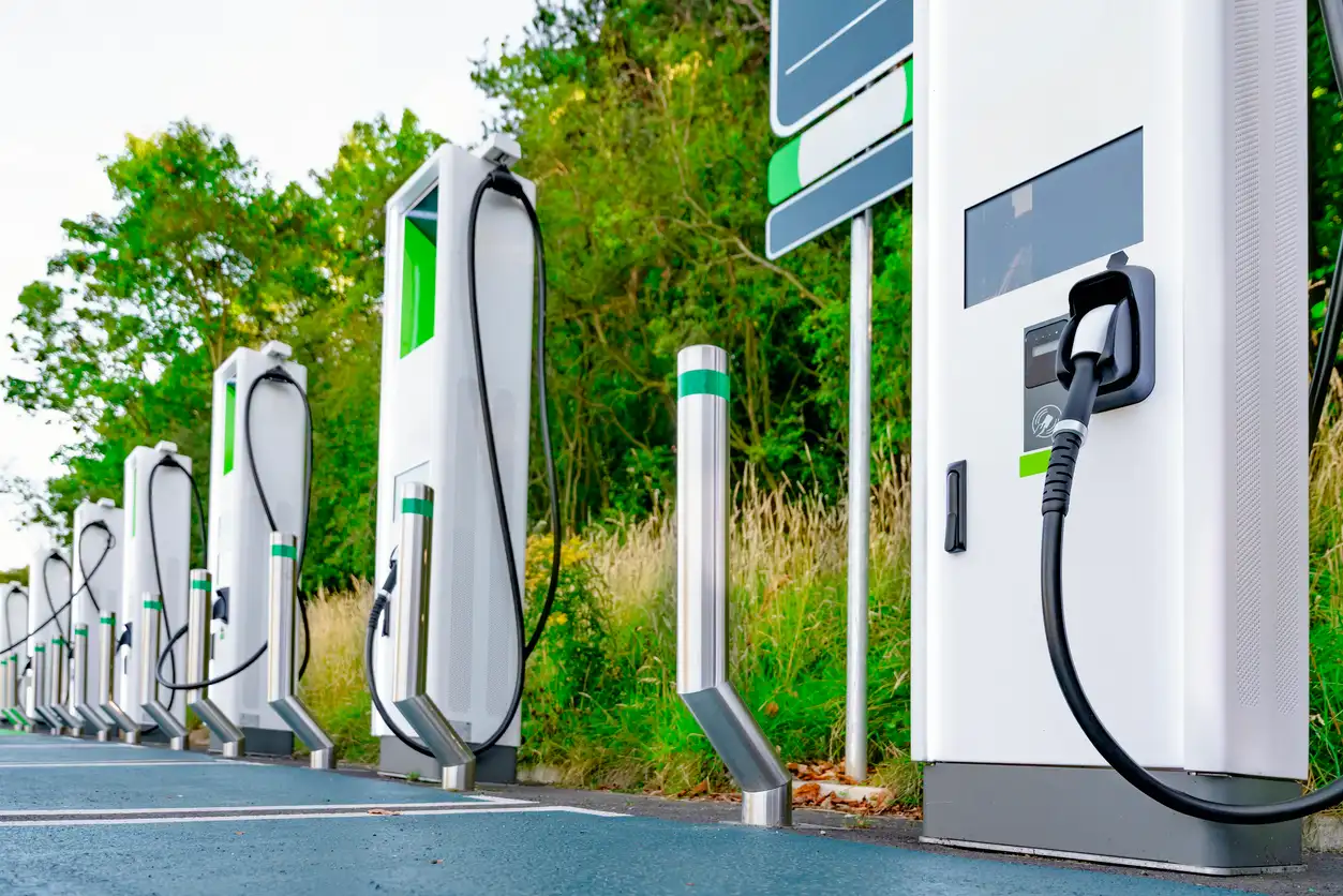 Postos de recarga para carros elétricos com conectores e cabos, em área externa ao lado de um sistema de energia, com vegetação ao fundo.