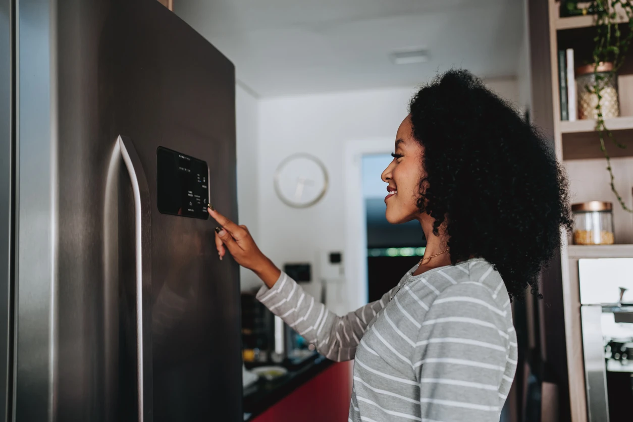 Mulher com cabelo cacheado usando o celular para acessar um painel na porta da geladeira inteligente em uma cozinha moderna, enquanto sorri.